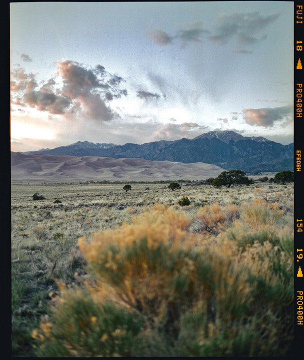 A vast landscape features a desert plain with shrubs in the foreground, sand dunes in the middle ground, and mountains under a cloudy sky.