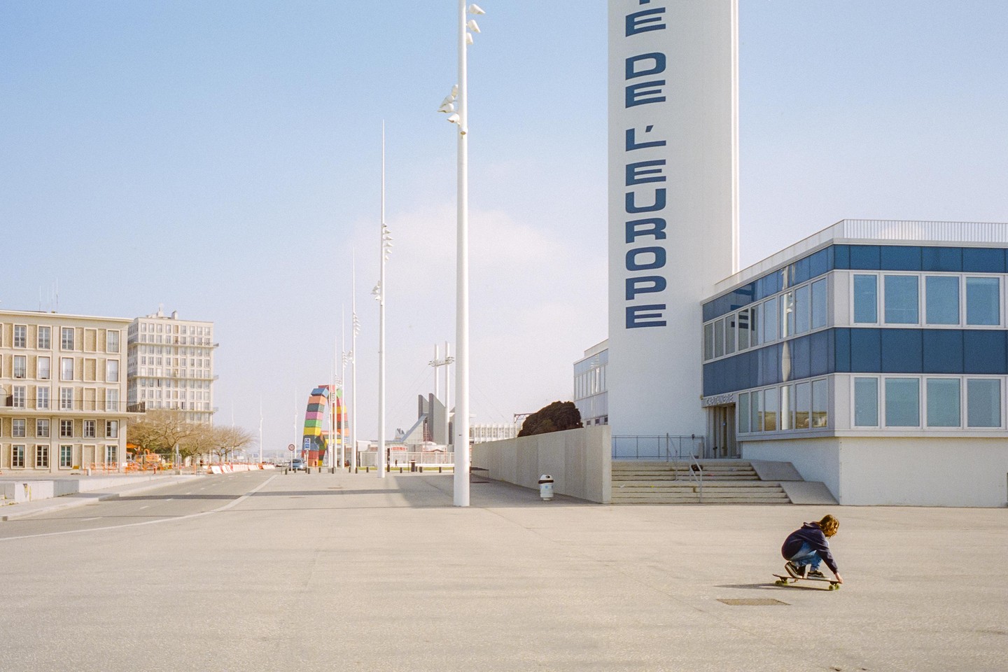 A child on a skateboard is in the foreground of a street scene with buildings and a colorful archway in the background.