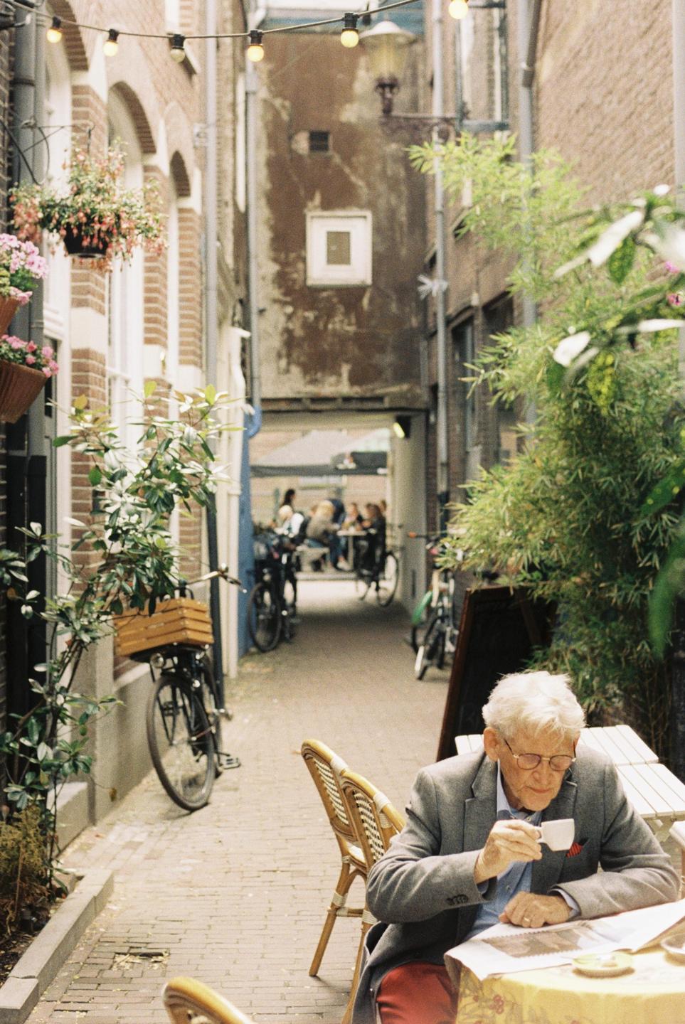 An elderly man in a blazer and red trousers sips coffee at an outdoor cafe table in a narrow alleyway.