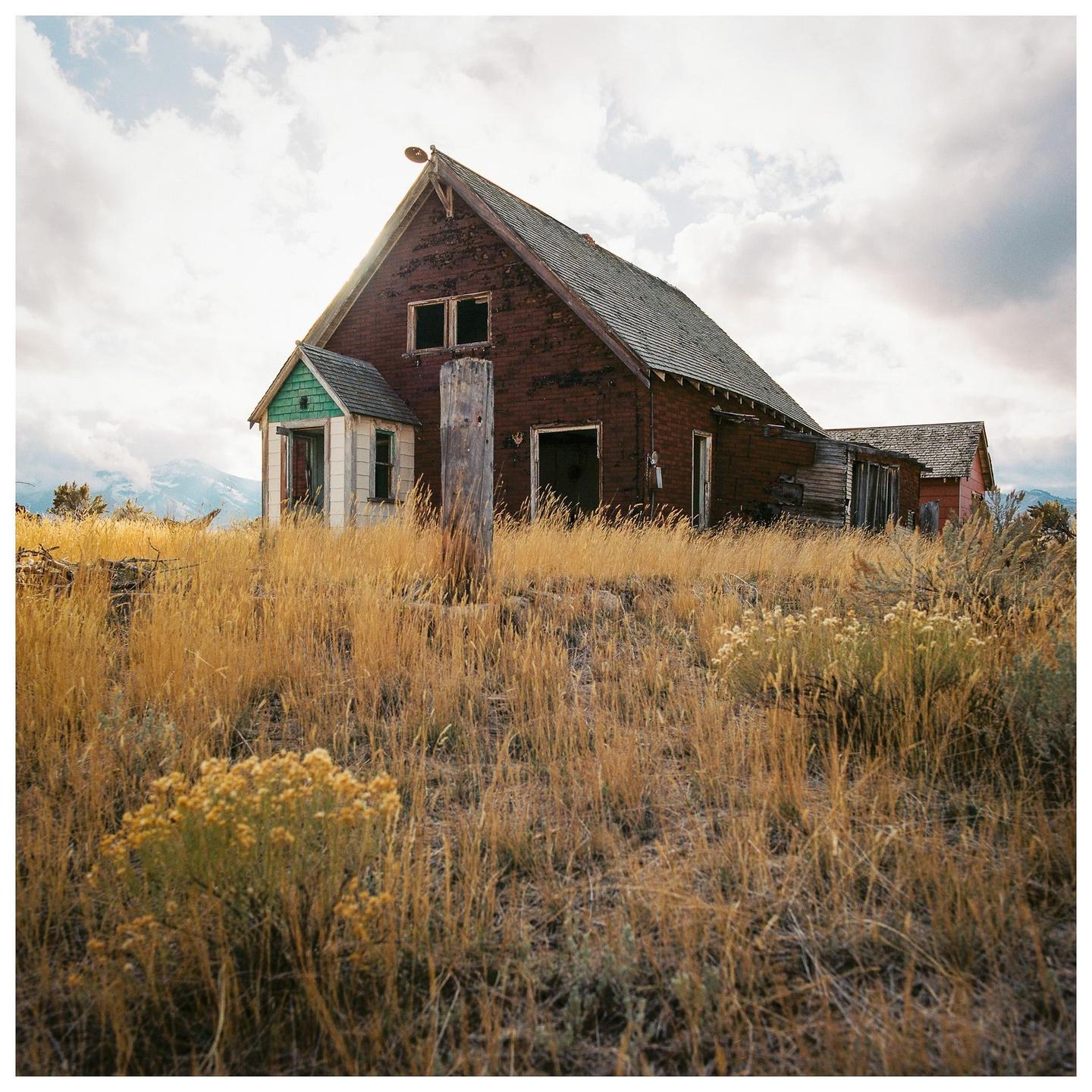 An old, weathered house with a green-painted porch stands in a field of tall, dry grass under a cloudy sky.