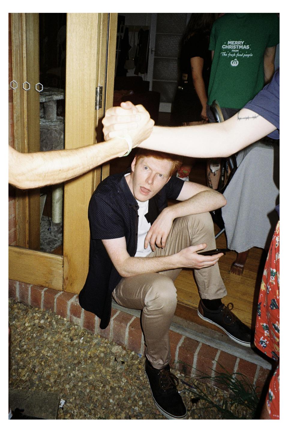 A man with red hair sits on a brick ledge while two arms shake hands above his head.