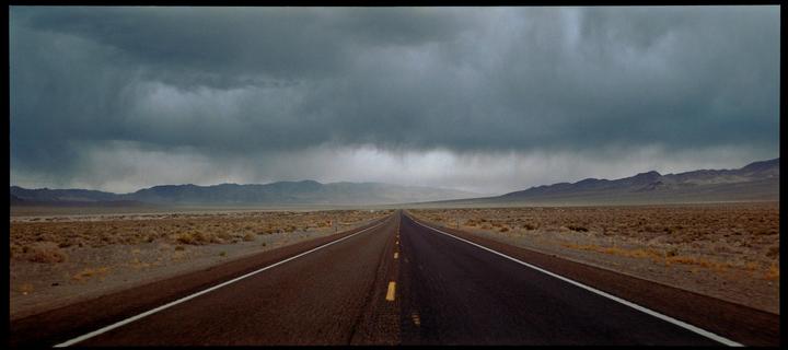 A long, straight road stretches through a desert landscape under a dramatic, cloudy sky.