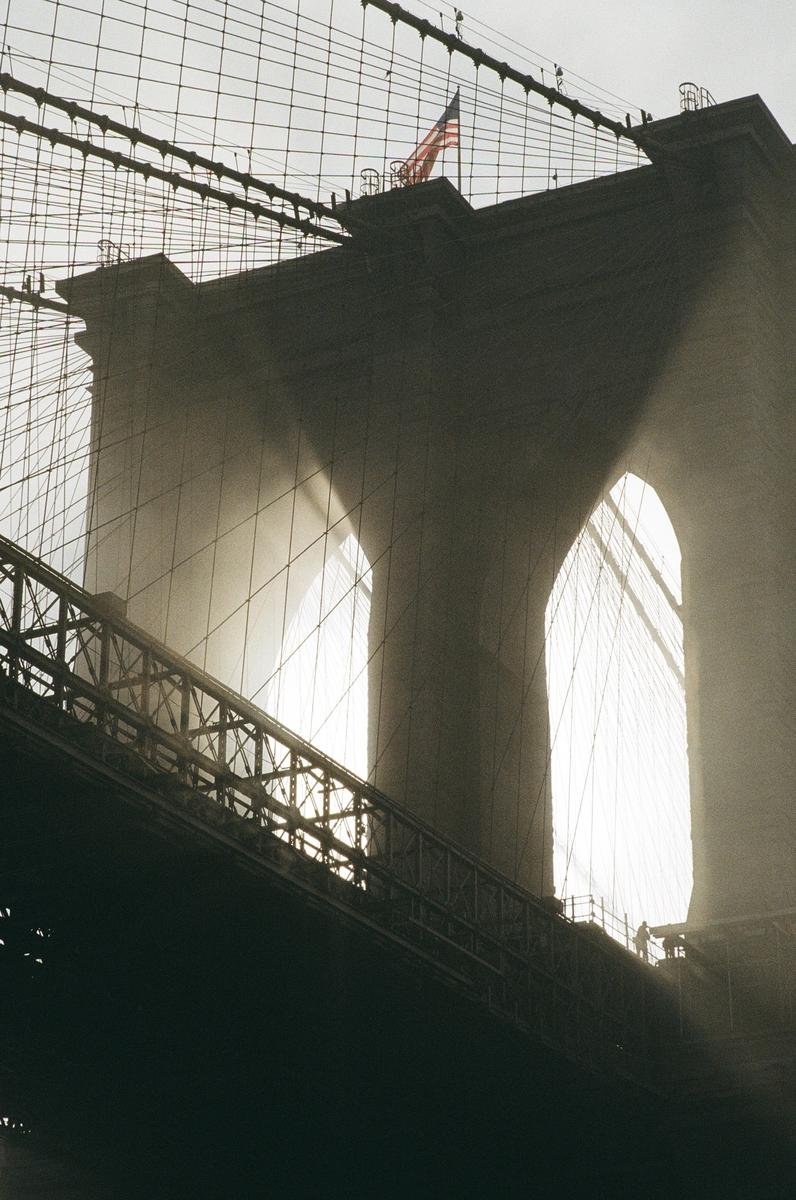 A low-angle view shows the Brooklyn Bridge tower and cables with a hazy, backlit sky.