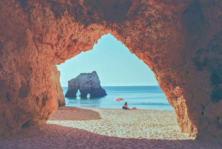 A woman sits on a sandy beach under a small umbrella, framed by a rock archway, with a large rock formation in the ocean.
