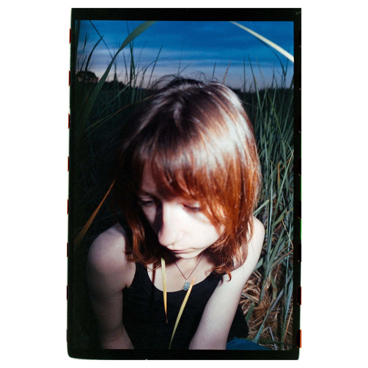A woman with red hair looks down while sitting in tall grass at dusk.