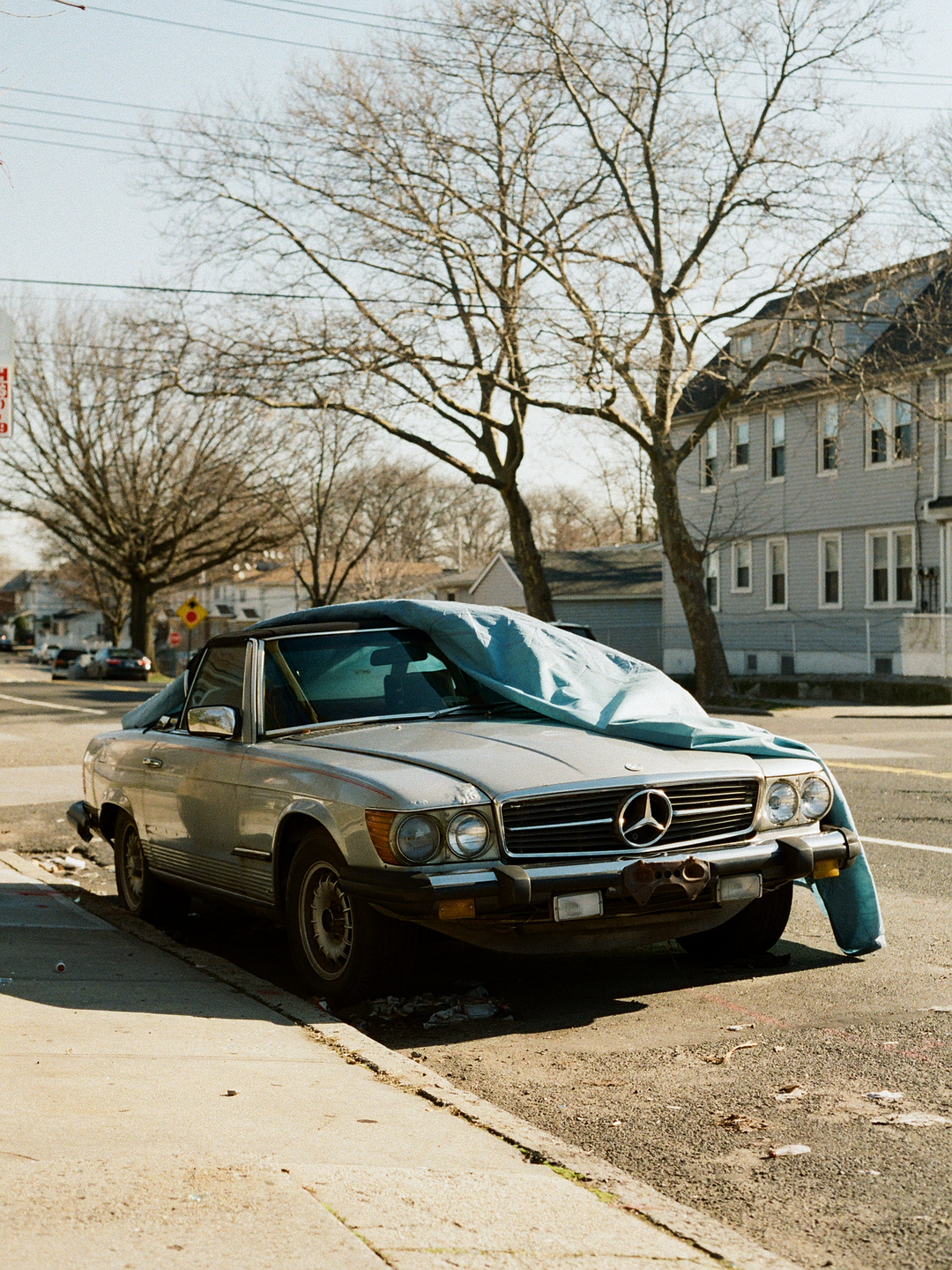 A silver convertible car with a blue cover on its hood is parked on a street next to a sidewalk, with bare trees and buildings in the background.