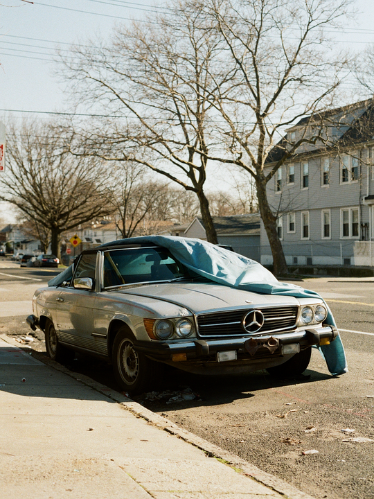 A silver convertible car with a blue cover on its hood is parked on a street next to a sidewalk, with bare trees and buildings in the background.