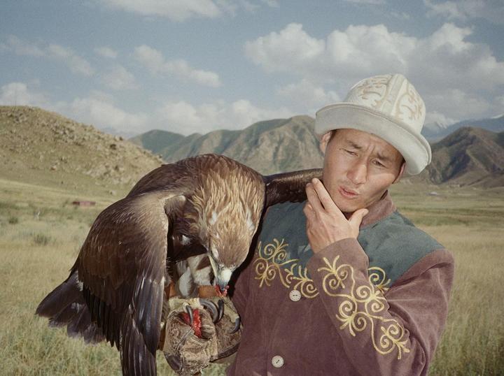 A man in traditional clothing holds an eagle on his arm in a mountainous, grassy landscape.