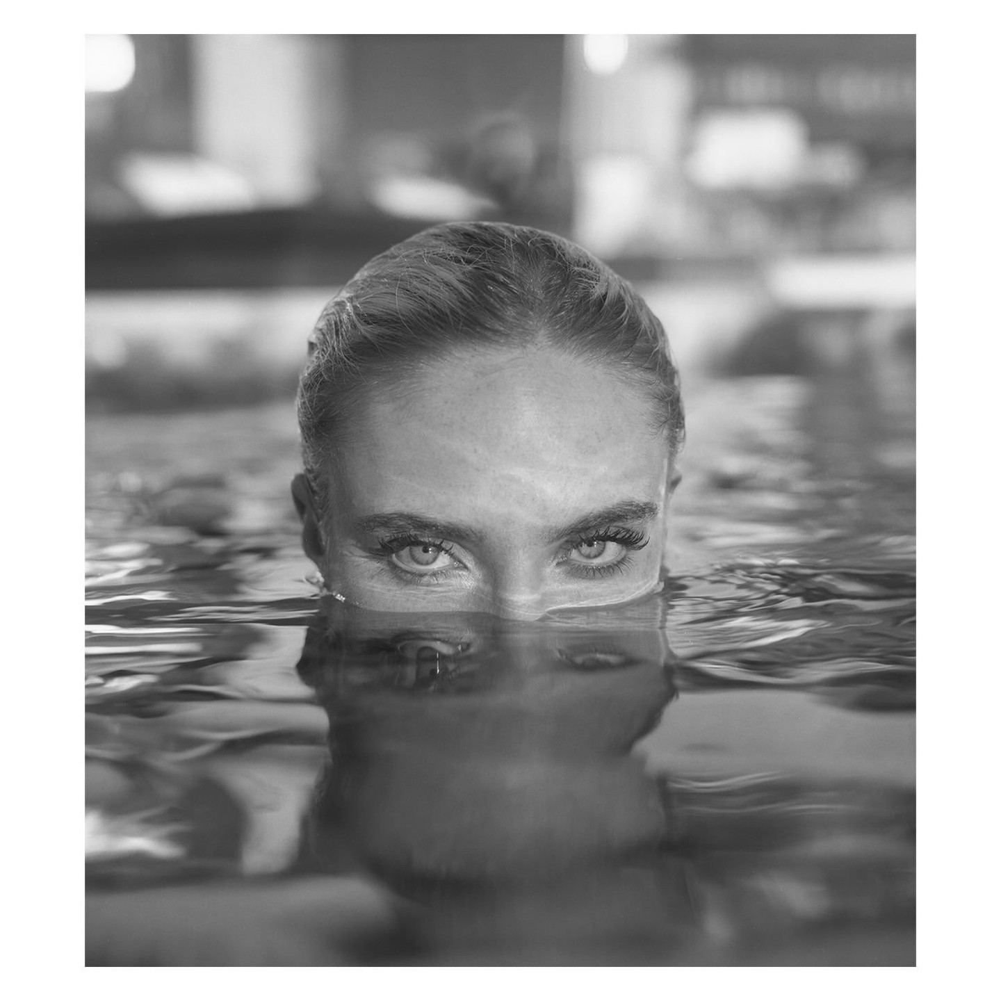 A woman's face is reflected in the water as she looks directly at the viewer.