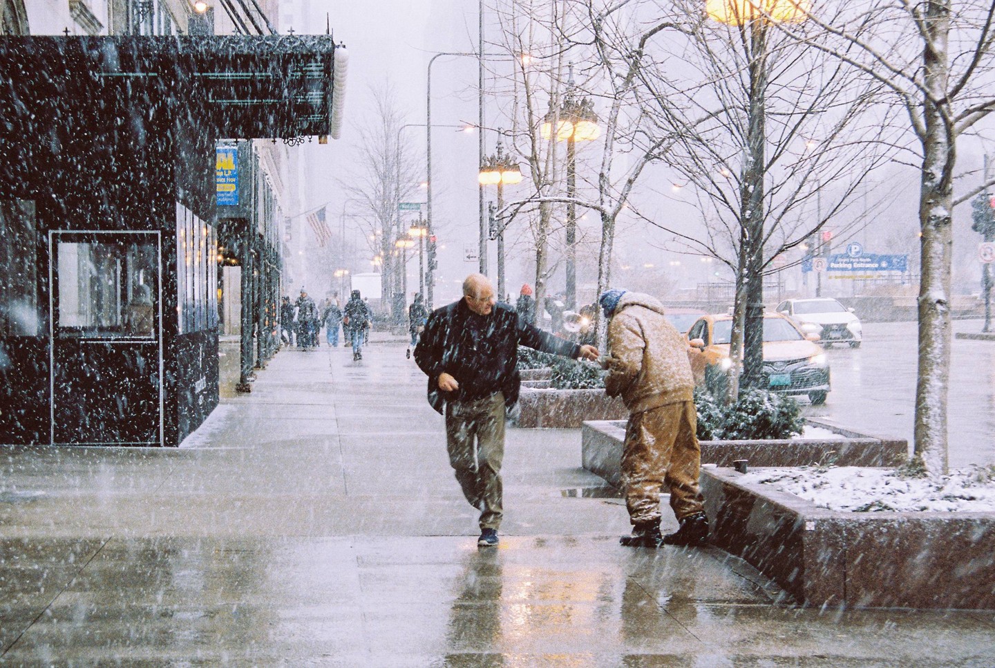 A man in a dark jacket reaches out to another man in a tan jacket on a snowy city street.