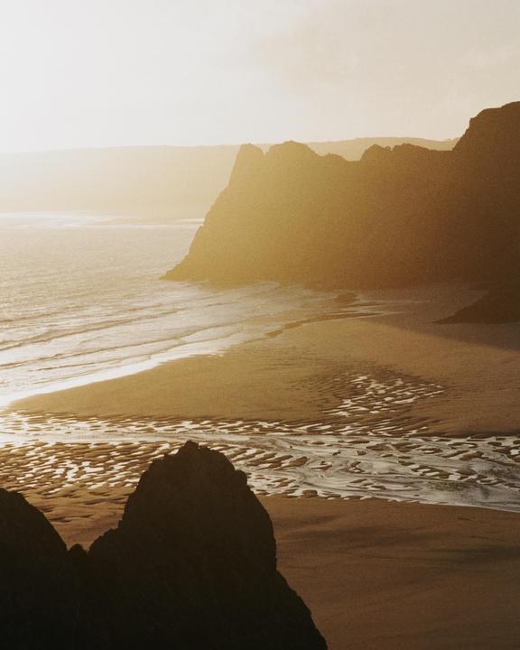 The sun shines over a beach with cliffs and ocean waves.