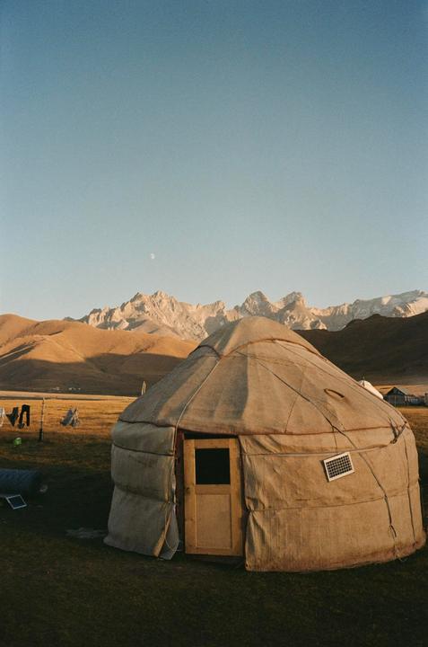 A yurt stands in a grassy field with mountains and a moon in the background.