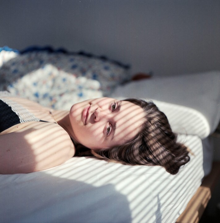 A woman lies on a bed with striped shadows cast across her face and the sheets.