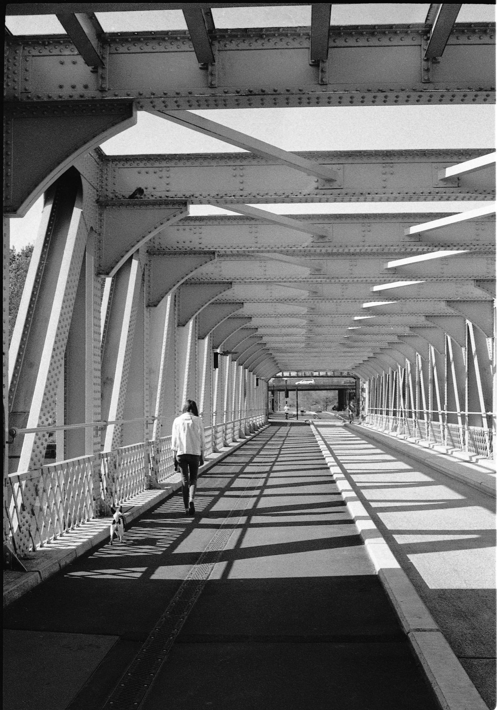A woman walks a small dog across a metal bridge with strong shadows cast by the structure.