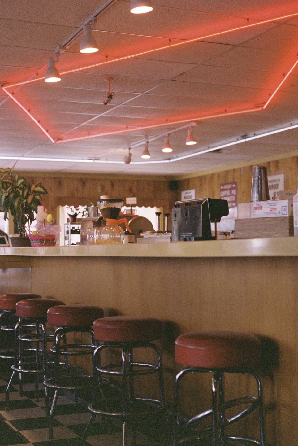 A diner interior features a counter with several red-topped stools and a neon light fixture on the ceiling.