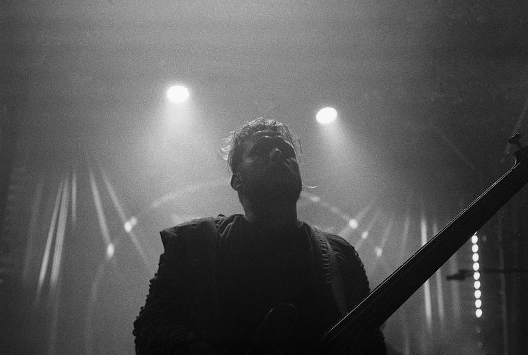 A man with messy hair is playing a bass guitar on a stage with bright spotlights.