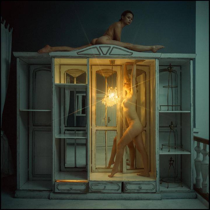 A nude woman sits in a split on top of a large dollhouse while another nude woman stands inside the dollhouse next to a glowing chandelier.