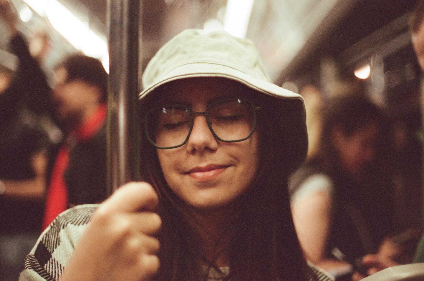 A woman wearing a bucket hat and glasses smiles with her eyes closed while holding onto a pole on a moving train.