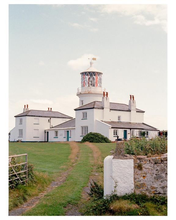 A white lighthouse and buildings stand on a grassy hill with a dirt path leading up to them.