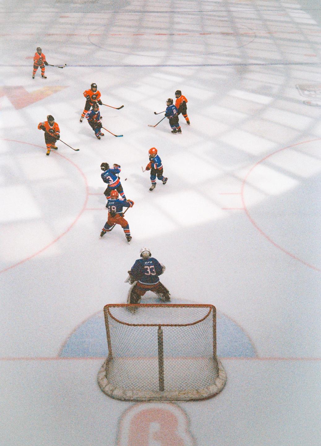 A high-angle view shows a hockey game in progress with players in orange and blue jerseys on an ice rink.