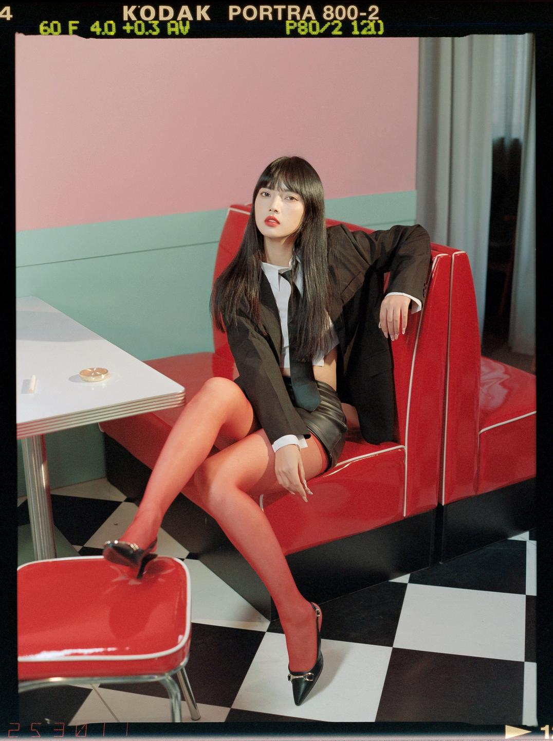 A woman in a black blazer, red tights, and a black skirt sits on a red diner booth next to a white table.