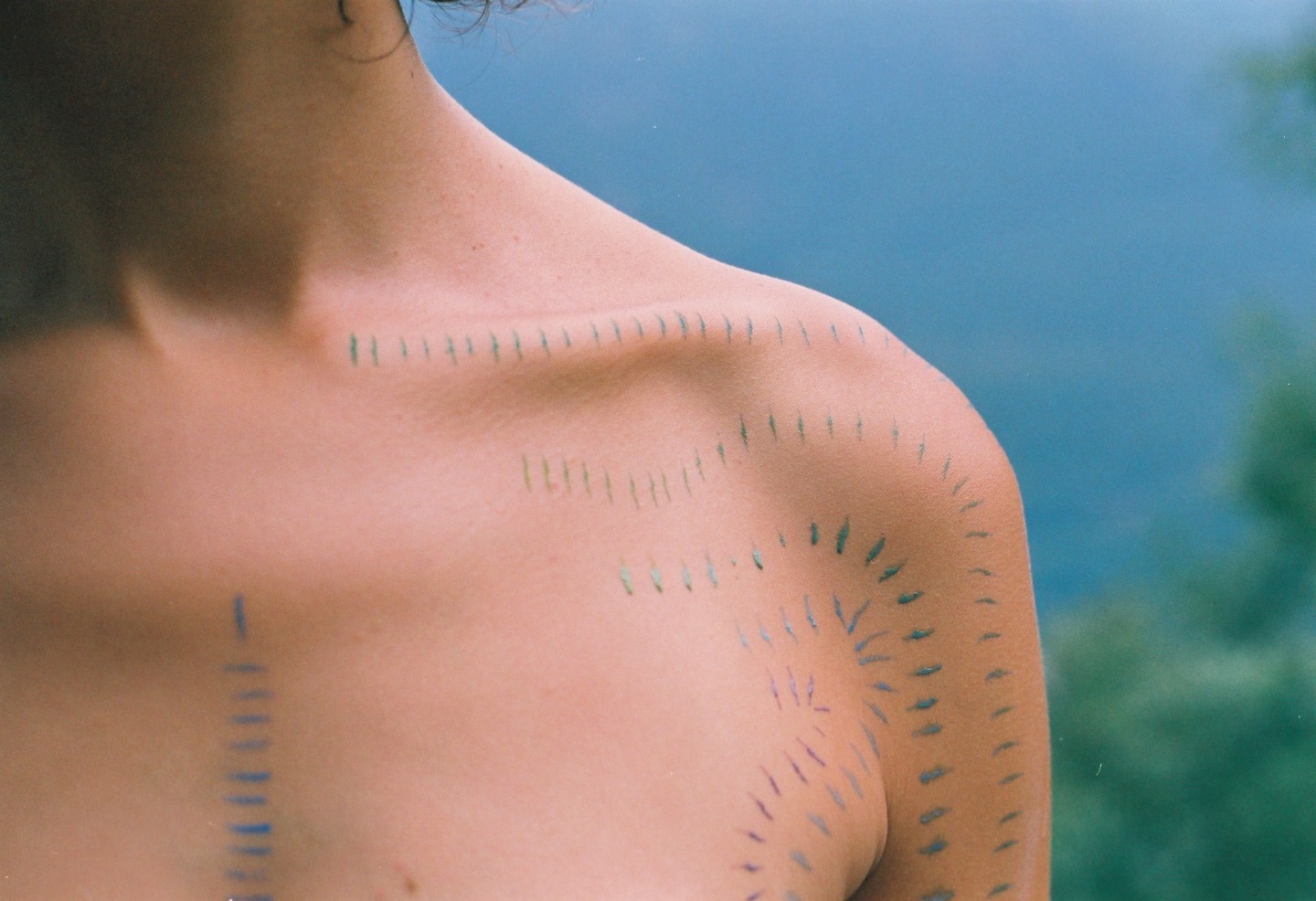 A person's shoulder and chest are marked with small, painted lines against a blue and green background.
