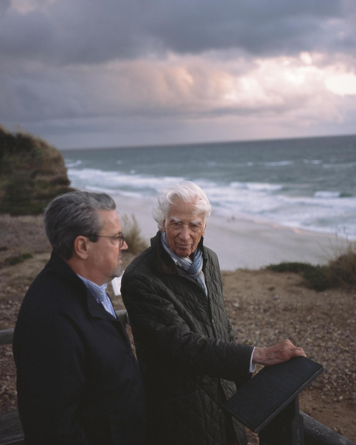 Two men stand on a cliff overlooking a beach and ocean under a cloudy sky.