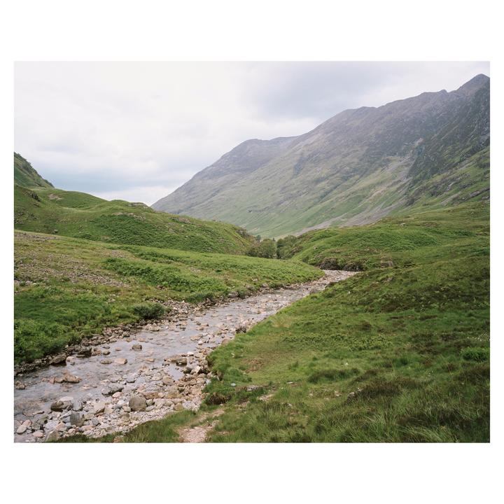 A rocky stream flows through a green valley with mountains under a cloudy sky.