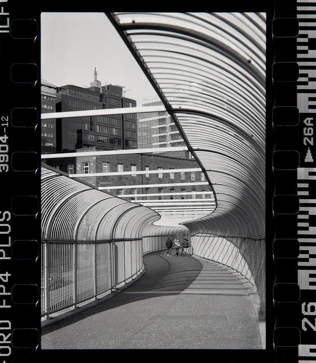 Three people walk through a curved, covered walkway with buildings in the background.