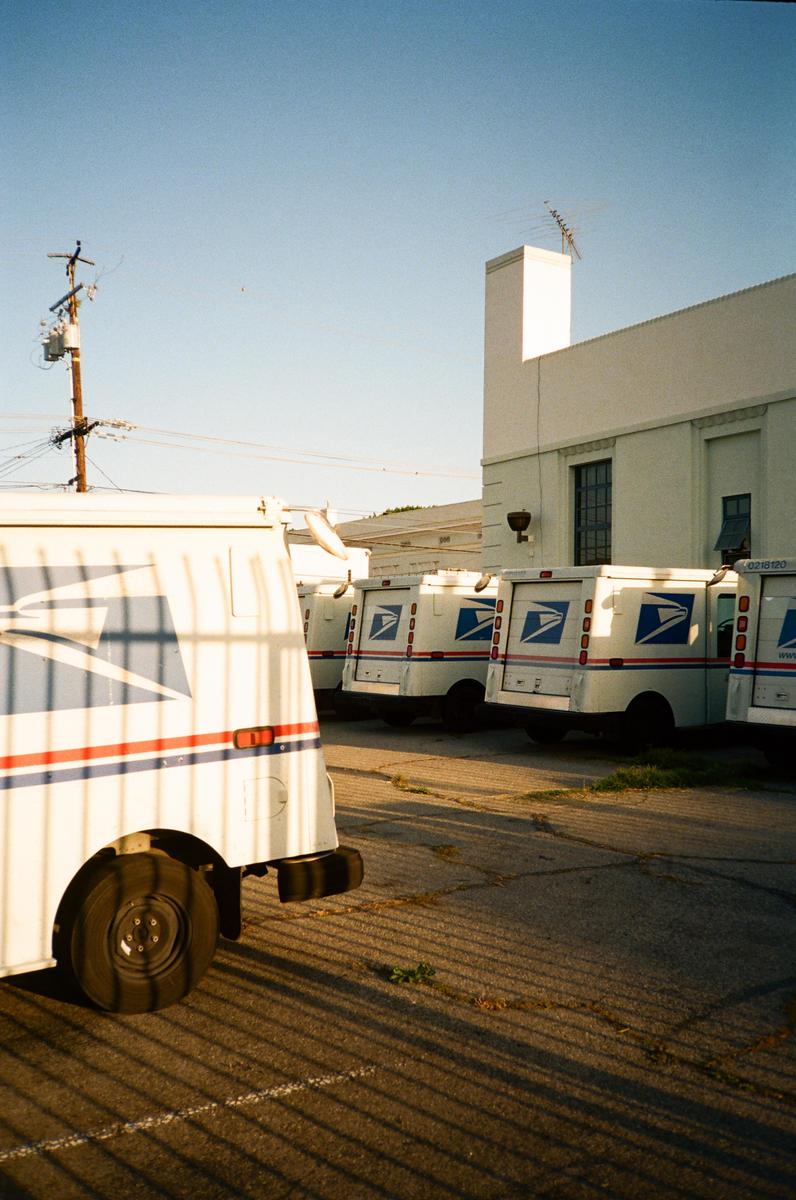 Several white postal vans are parked in a lot next to a building under a clear sky.
