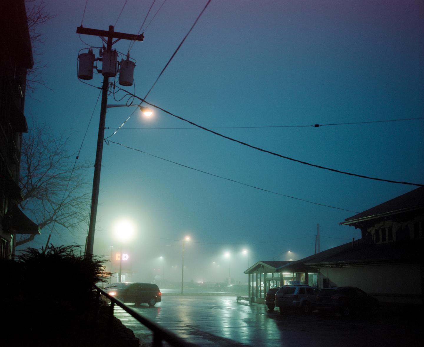 A street at night is illuminated by streetlights and car lights, with a utility pole and wires in the foreground.