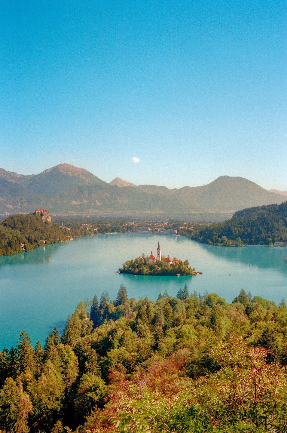 A small island with a church sits in the middle of a turquoise lake, surrounded by mountains and forests under a clear blue sky.