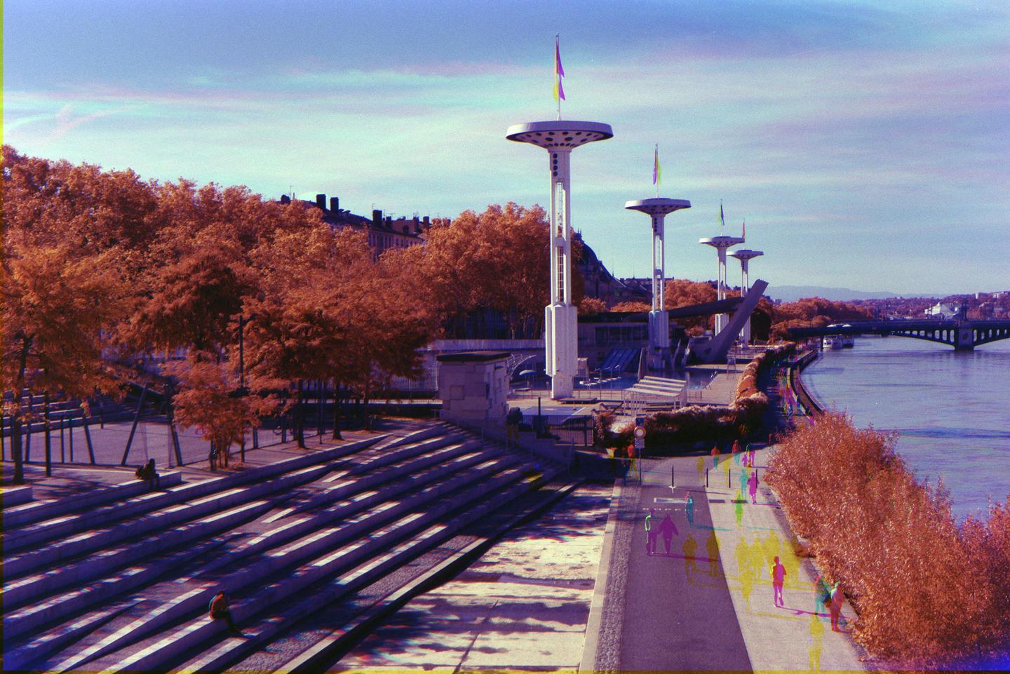 A wide view shows a river, a bridge, a promenade, and a large set of steps, all lined with trees and modern light fixtures.