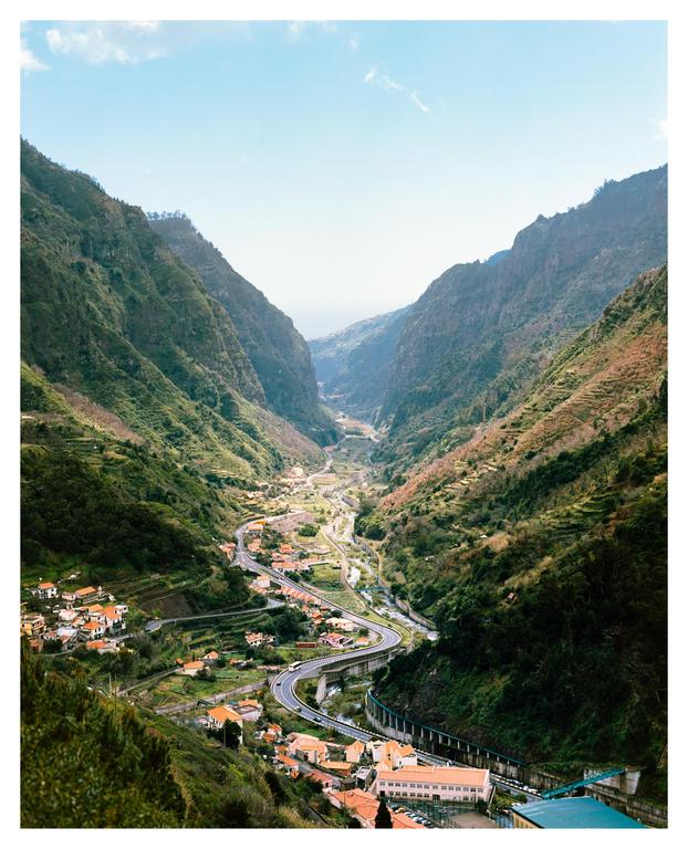 A winding road and a small village are nestled in a deep valley surrounded by lush green mountains under a clear sky.