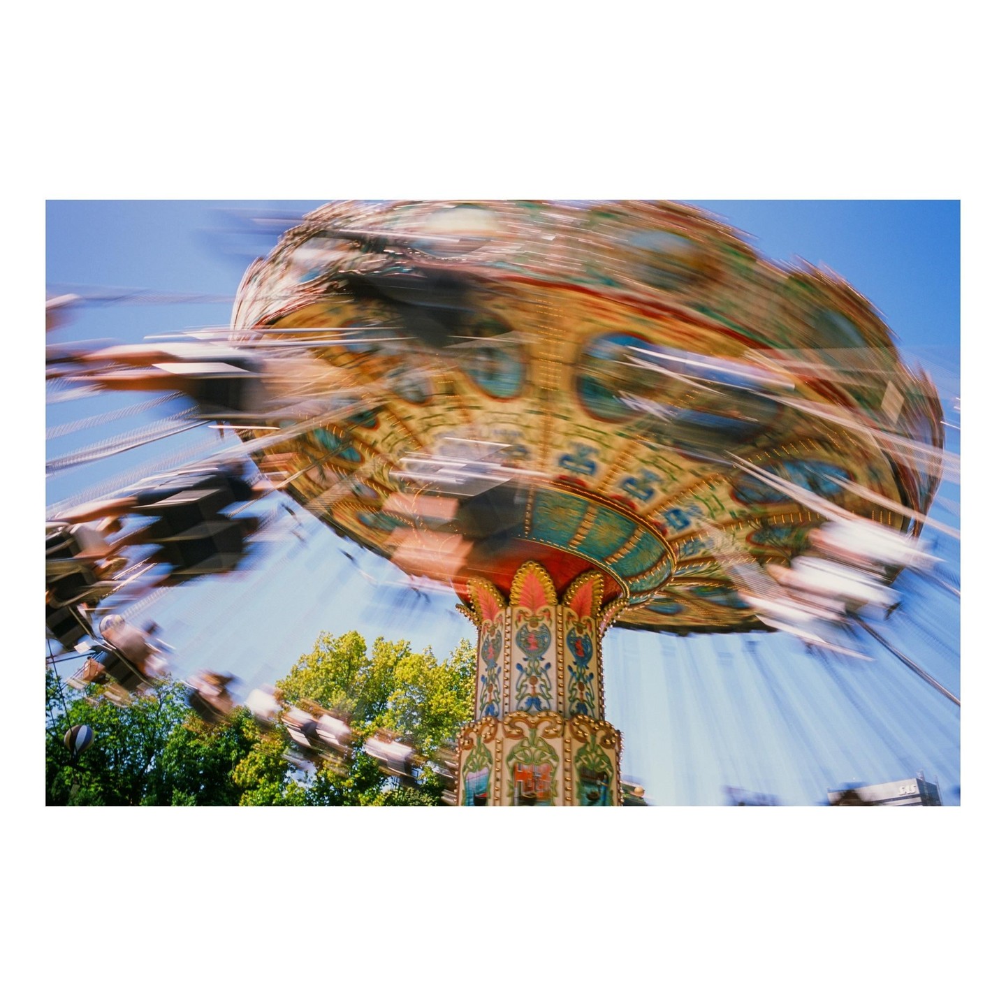 A colorful carousel ride spins against a clear blue sky with green trees in the background.