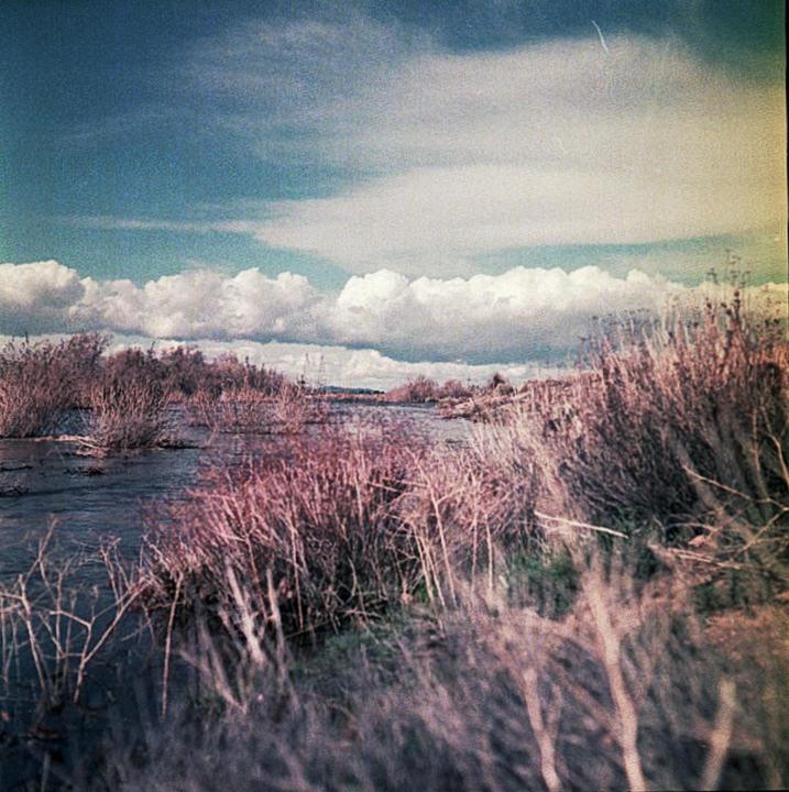 A river flows through a landscape with dry brush and a cloudy sky.