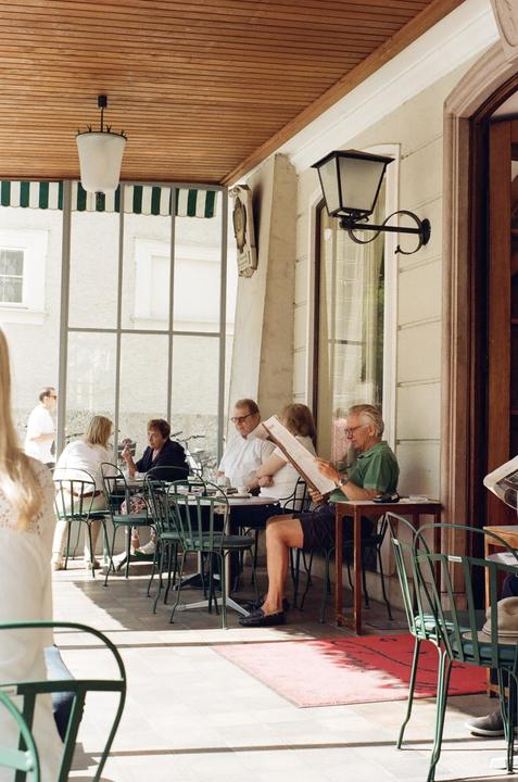 People are seated at tables on a covered patio outside a building with large windows.