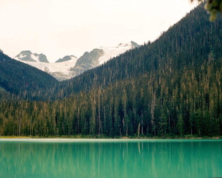 A turquoise lake reflects a dense forest and snow-capped mountains under a hazy sky.