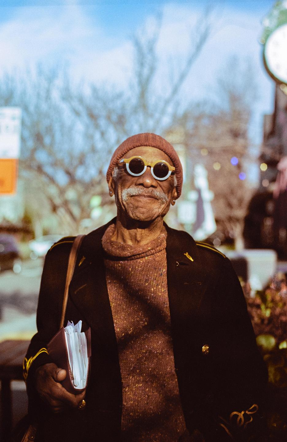 A man wearing a beanie and sunglasses smiles at the camera while holding a small book.