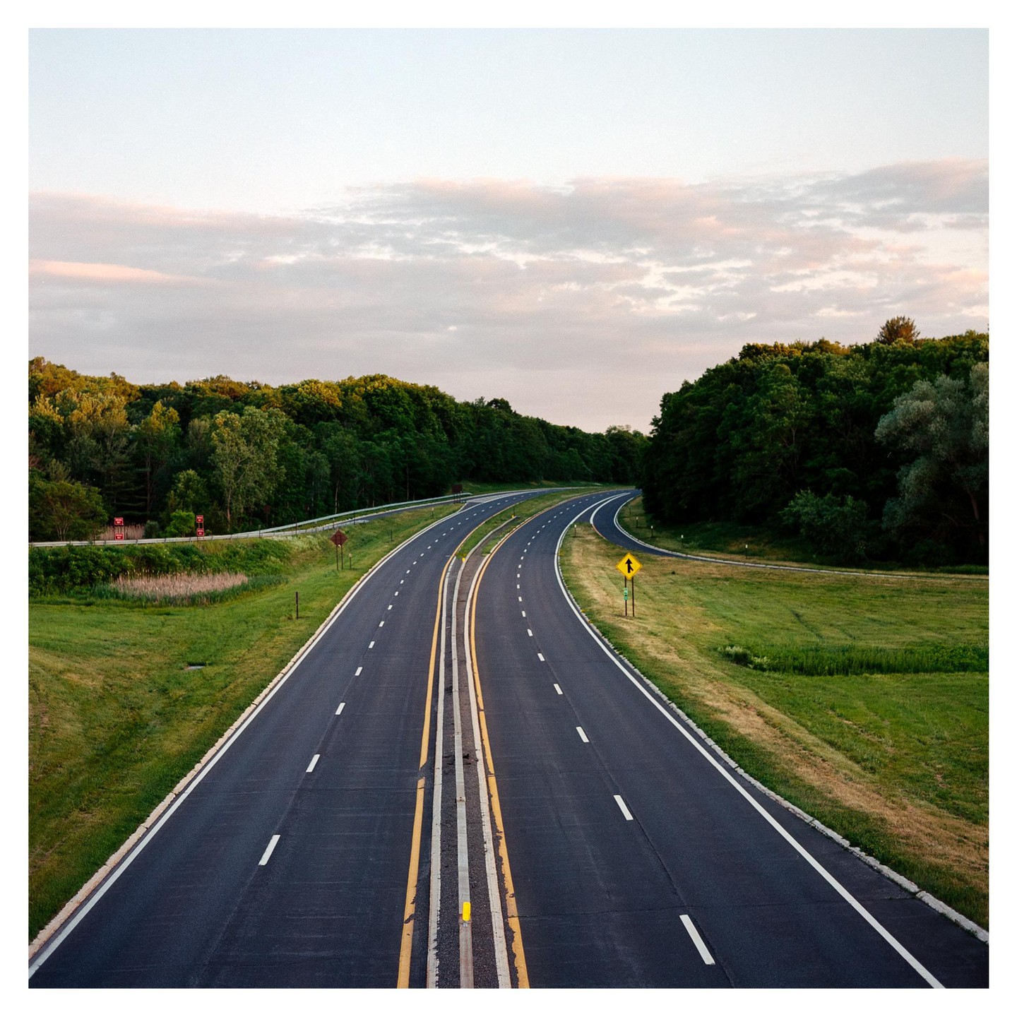 A highway with a grassy median and surrounding trees curves under a cloudy sky.