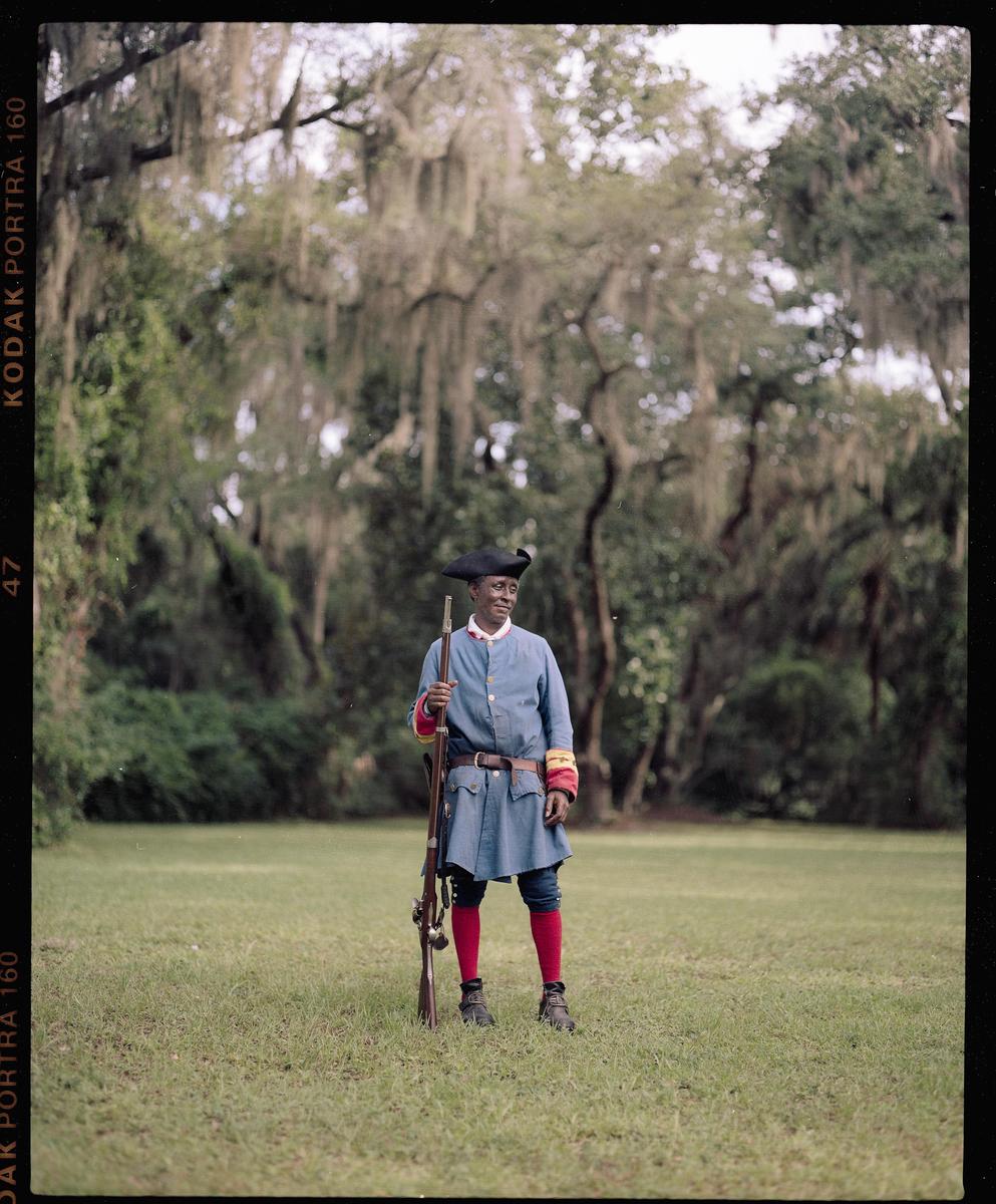A man in historical military attire stands in a grassy field holding a musket, with trees draped in spanish moss in the background.