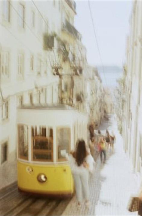 A woman stands next to a yellow tram on a blurry street with buildings and people in the background.