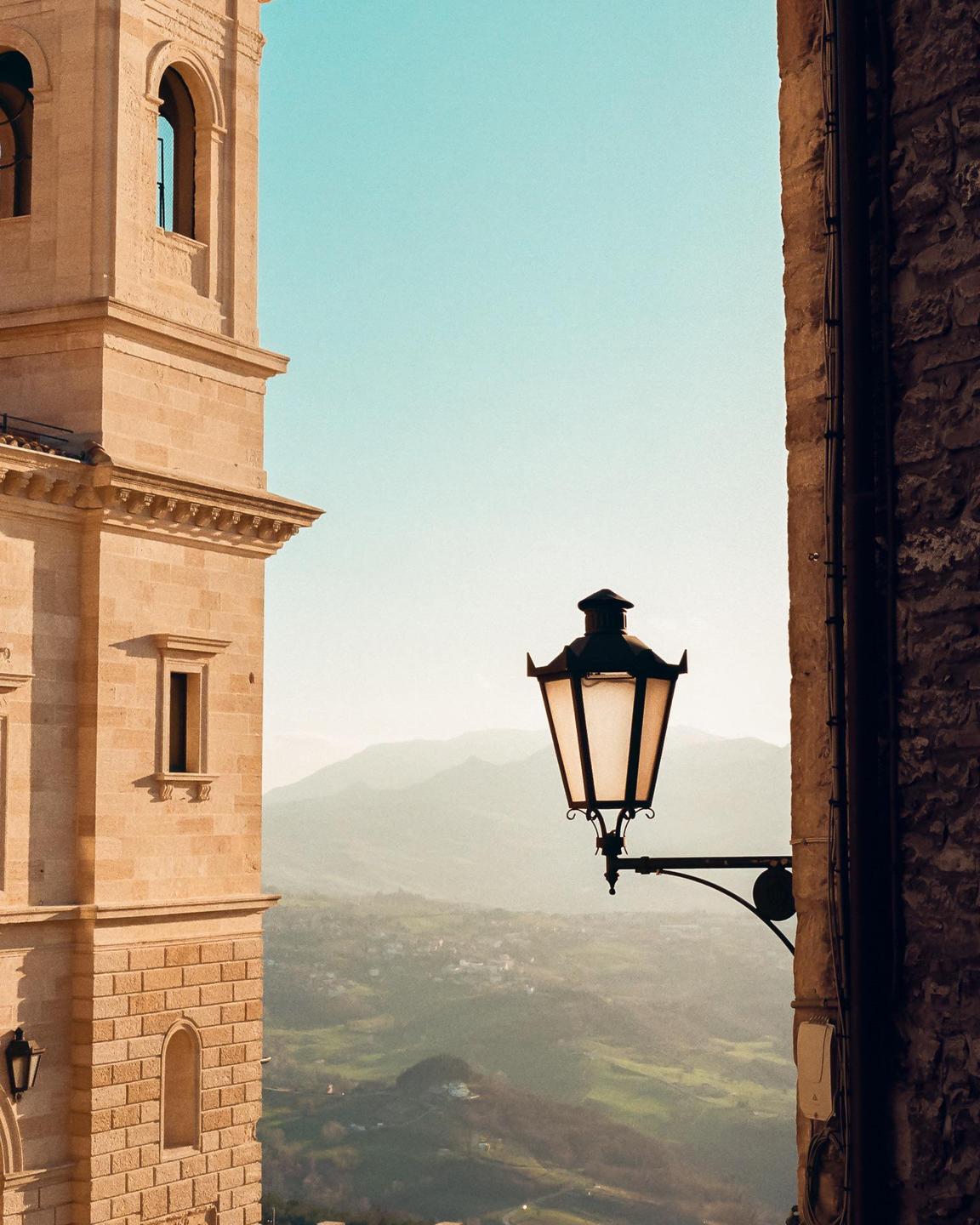 A street lamp is mounted on a stone wall with a view of mountains and a valley in the background.