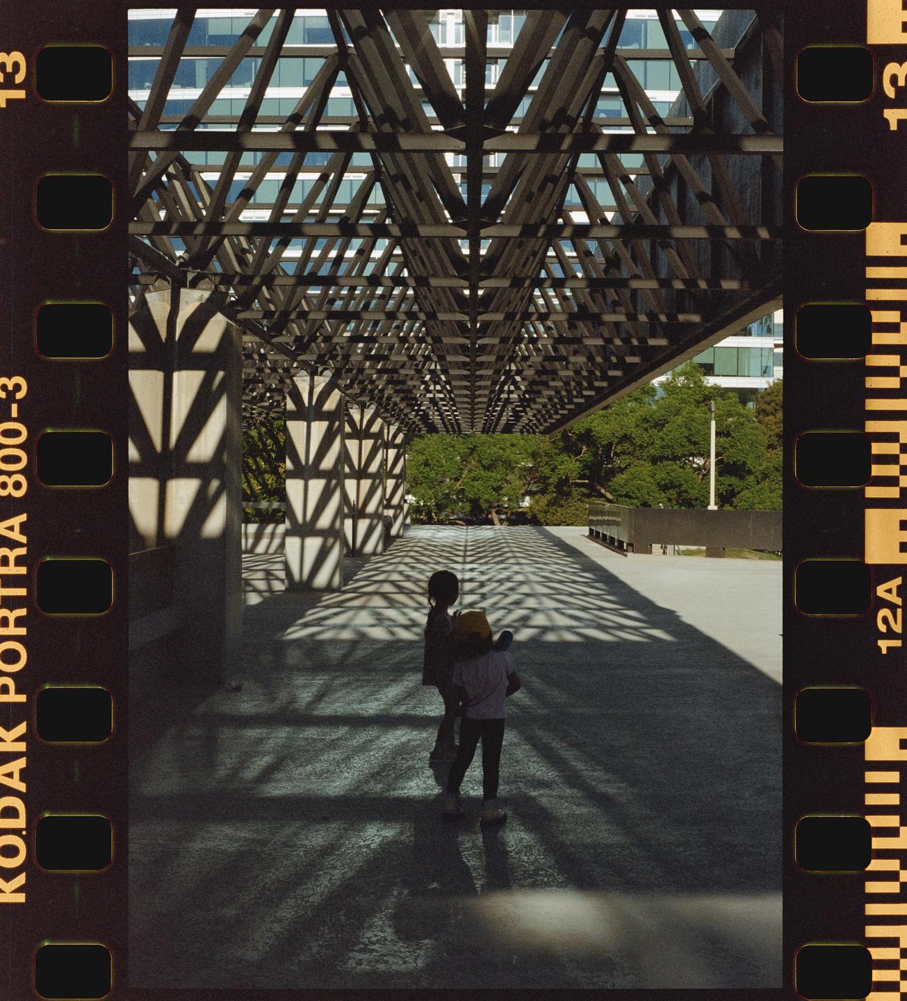 Two children stand on a patterned ground under a large, geometric structure with trees and a building in the background.