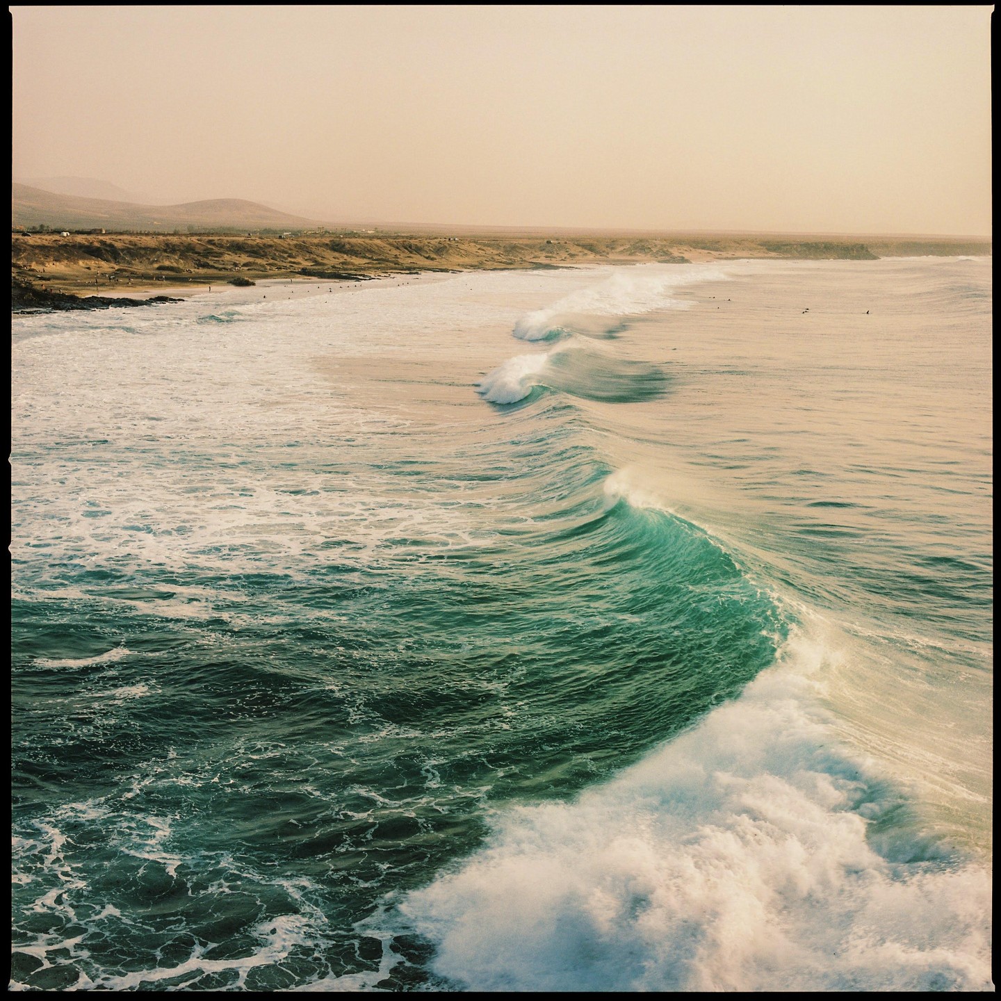 A large wave breaks on a beach with surfers in the distance under a hazy sky.