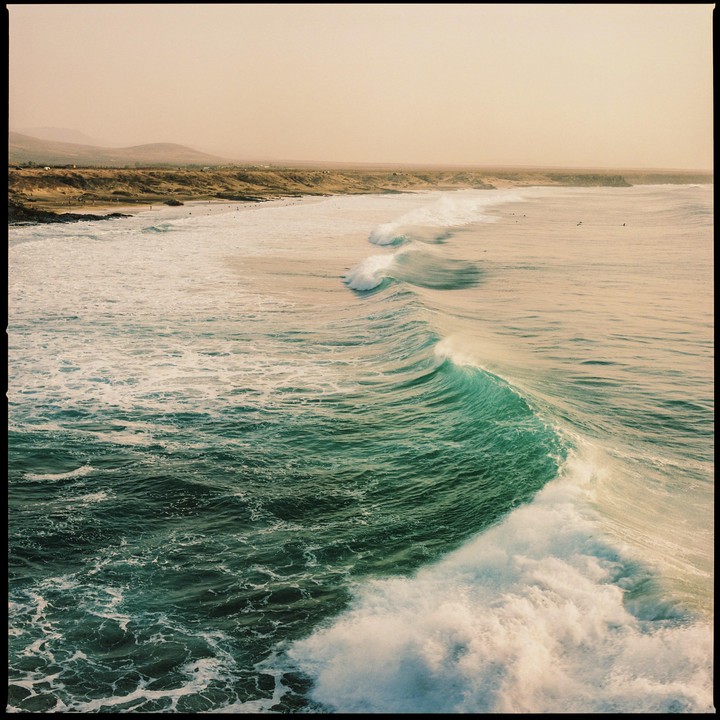 A large wave breaks on a beach with surfers in the distance under a hazy sky.