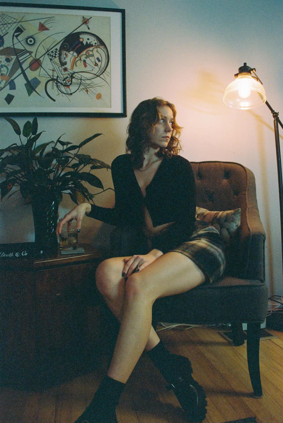 A woman sits in a chair next to a plant and a lamp, looking away from the camera.