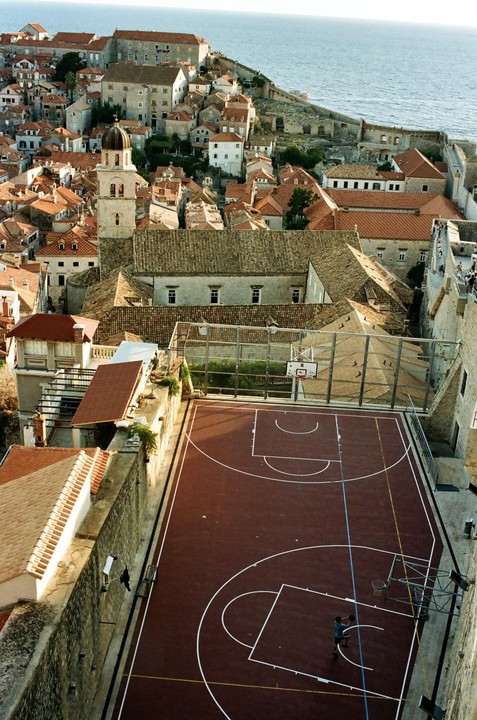 An aerial view shows a basketball court nestled among buildings with terracotta roofs, with the sea visible in the background.