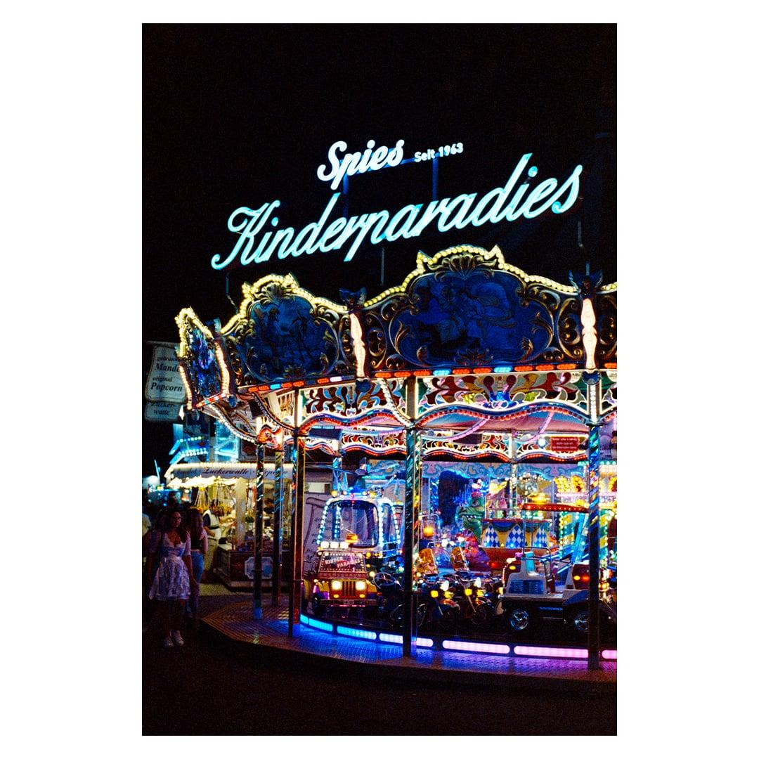 A brightly lit carousel with a neon sign that reads "Spies Kinderparadies" stands out against the dark night sky.