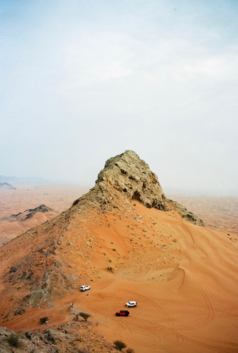 A rocky peak rises from a vast desert landscape with several vehicles on the sand dunes.
